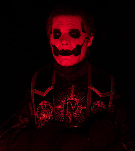 papa emeritus iv, wearing black and white skullpaint and a black alb with embroidered details and stole in front of a black background lit up by harsh red lighting. he holds his mic to his chest, his expression is neutral and his lips are parted slightly