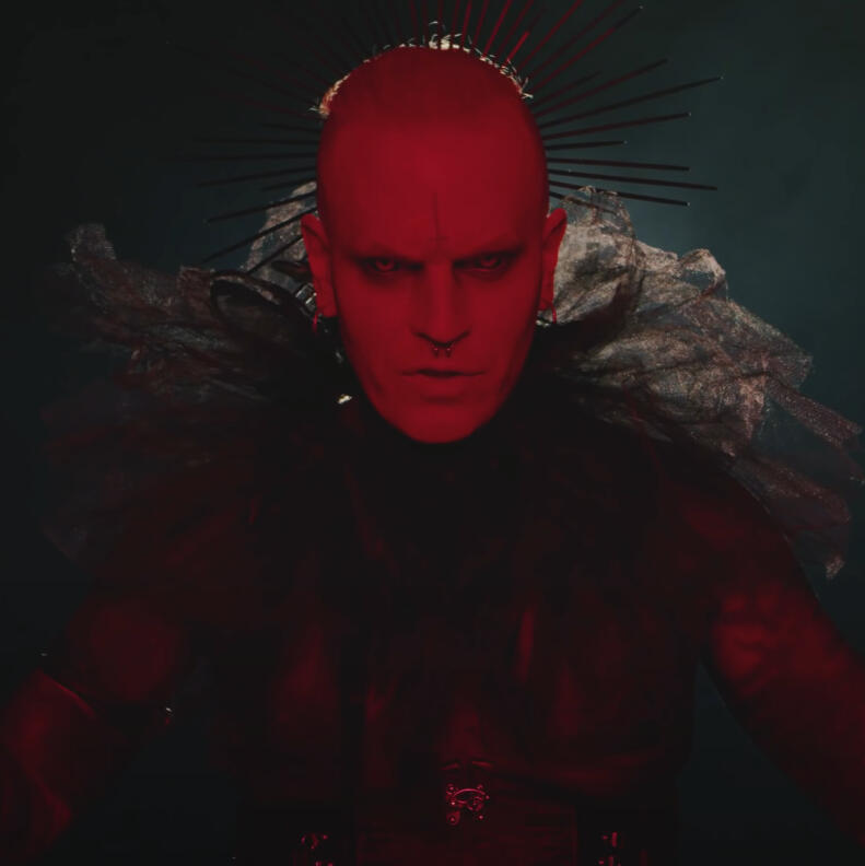 chris harms from lord of the lost in red light. he's wearing a black corset a tulle neckpiece and a sunburst crown. his body is smudged with black paint. he has an inverted j shaped cross on his forehead and red eye contacts. his ears and nose are pierced