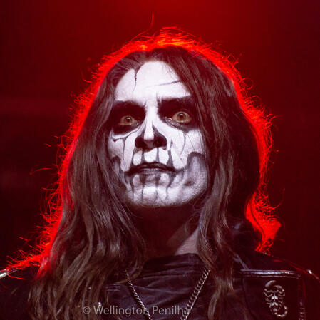 the vocalist seregor from carach angren in red lighting onstage. his corpse paint is ghastly black and white with contoured cheek bones. he has long wavy brown hair